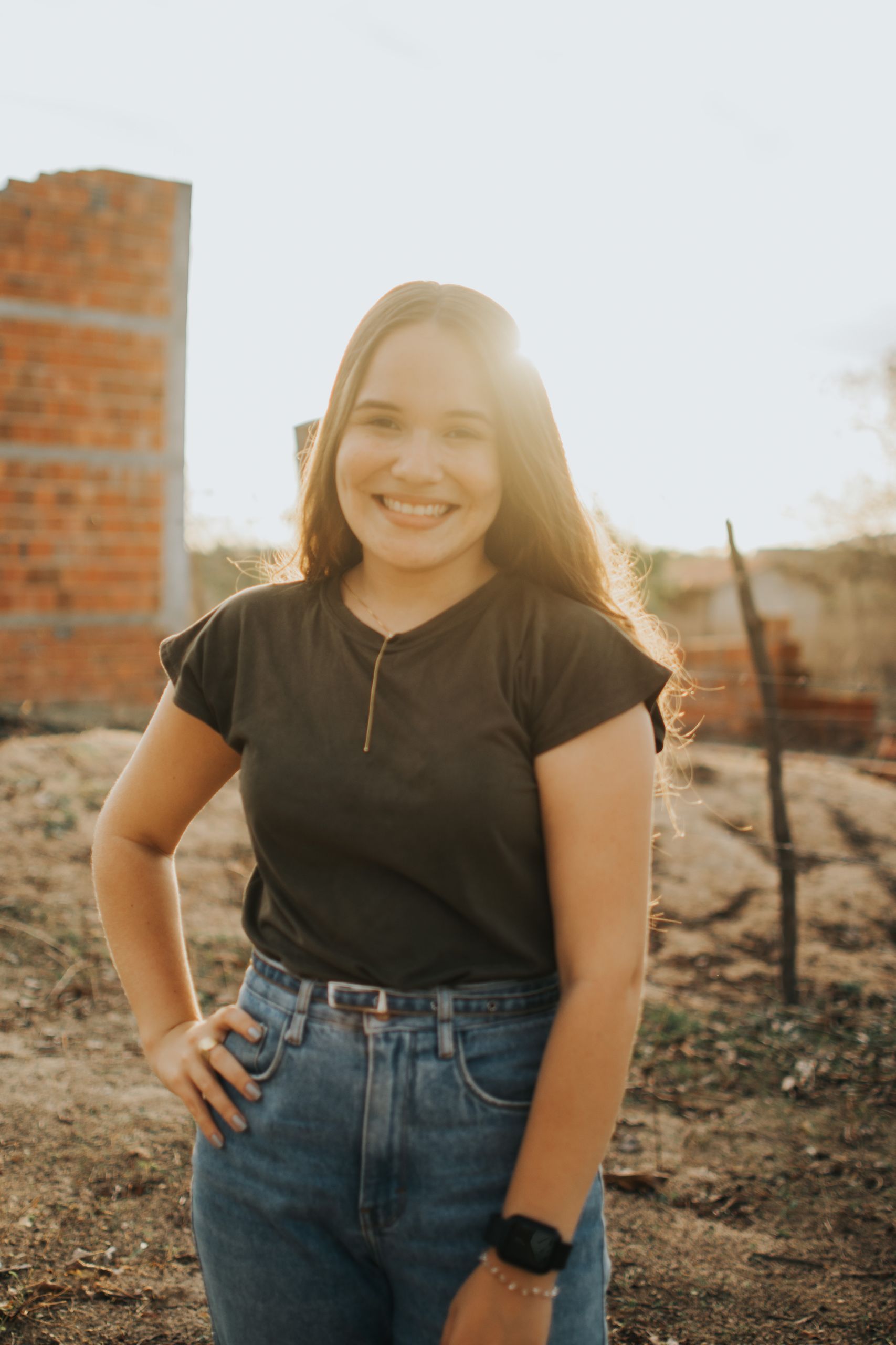 A cheerful young woman stands outdoors at sunset, backlit by warm sunlight.