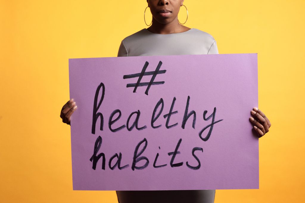 A photo of an adult woman holding a purple sign promoting healthy habits against a vibrant yellow background.