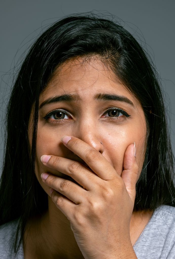 Close-up portrait of a woman with hand covering mouth, showing a scared expression, in a studio setting.